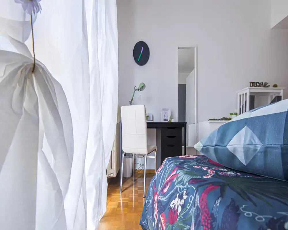 Angolo camera luminoso e curato con biancheria dai motivi decorativi e una scrivania; la luce naturale che filtra dalle tende crea un’atmosfera fresca e accogliente.