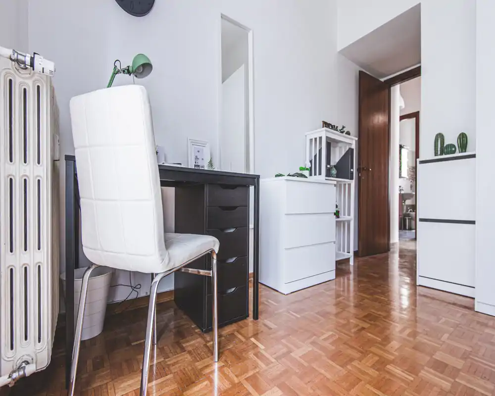 Zona giorno luminosa e minimal con una piccola scrivania e mobili contenitori bianchi; parquet in legno e linee pulite creano uno spazio funzionale ed ordinato.