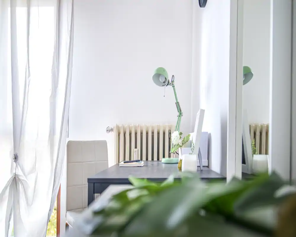 Angolo di lavoro luminoso con scrivania, lampada verde regolabile, sedia e pianta decorativa vicino a una finestra con tenda leggera — un angolo di lavoro arioso e minimale.