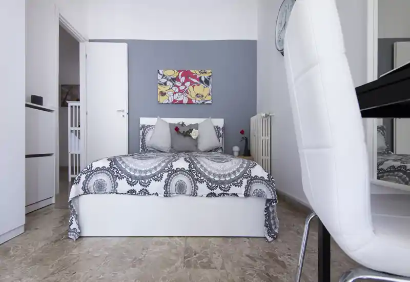 Bright modern bedroom with a centrally placed double bed, patterned duvet and decorative pillows; neutral grey and white palette with floral wall art and clean marble floor.