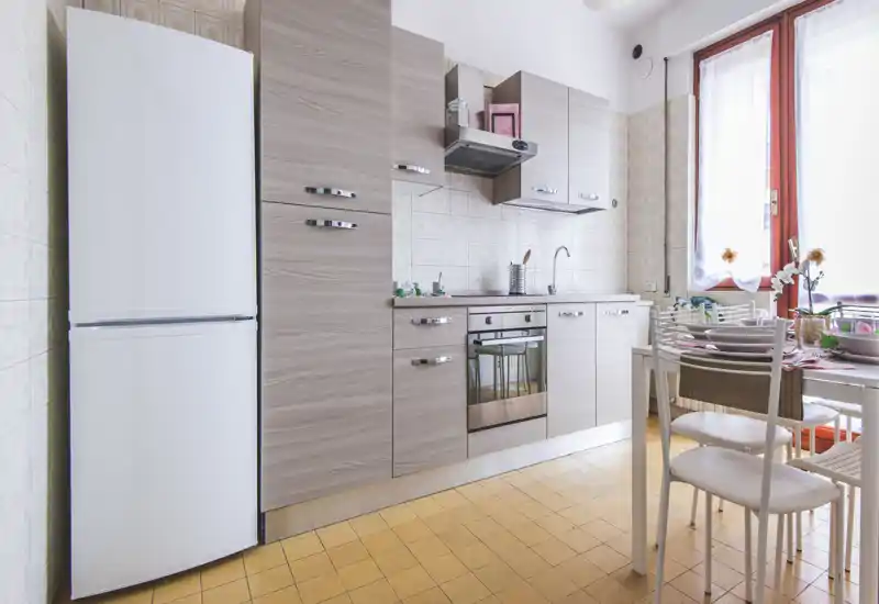 Cuisine lumineuse et moderne avec placards effet bois, four encastré, réfrigérateur et une petite table à manger près d'une fenêtre ensoleillée.