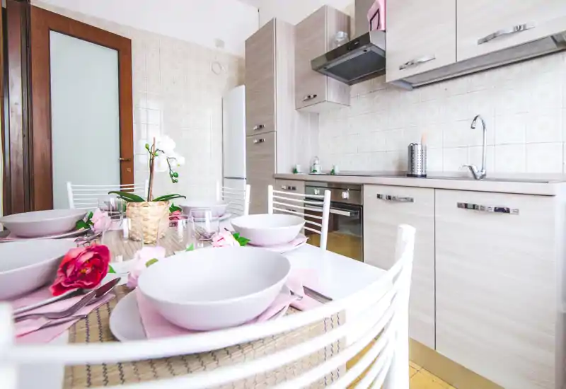 Cuisine moderne et lumineuse avec meubles en bois clair, électroménagers intégrés et table dressée avec accents roses et fleurs — un espace convivial pour des repas décontractés.