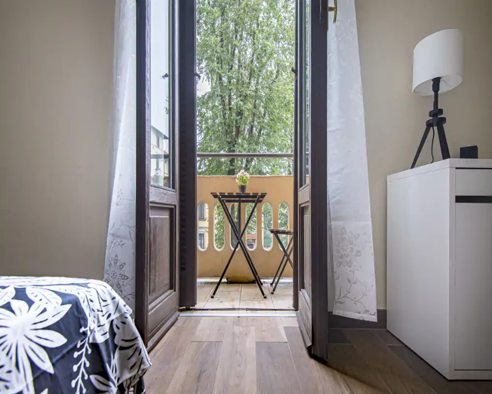 Camera da letto luminosa con accesso a un piccolo balcone e un tavolino bistro. Il letto ben rifatto, il comodino e la lampada creano un ambiente accogliente e ben illuminato, perfetto per gli annunci.