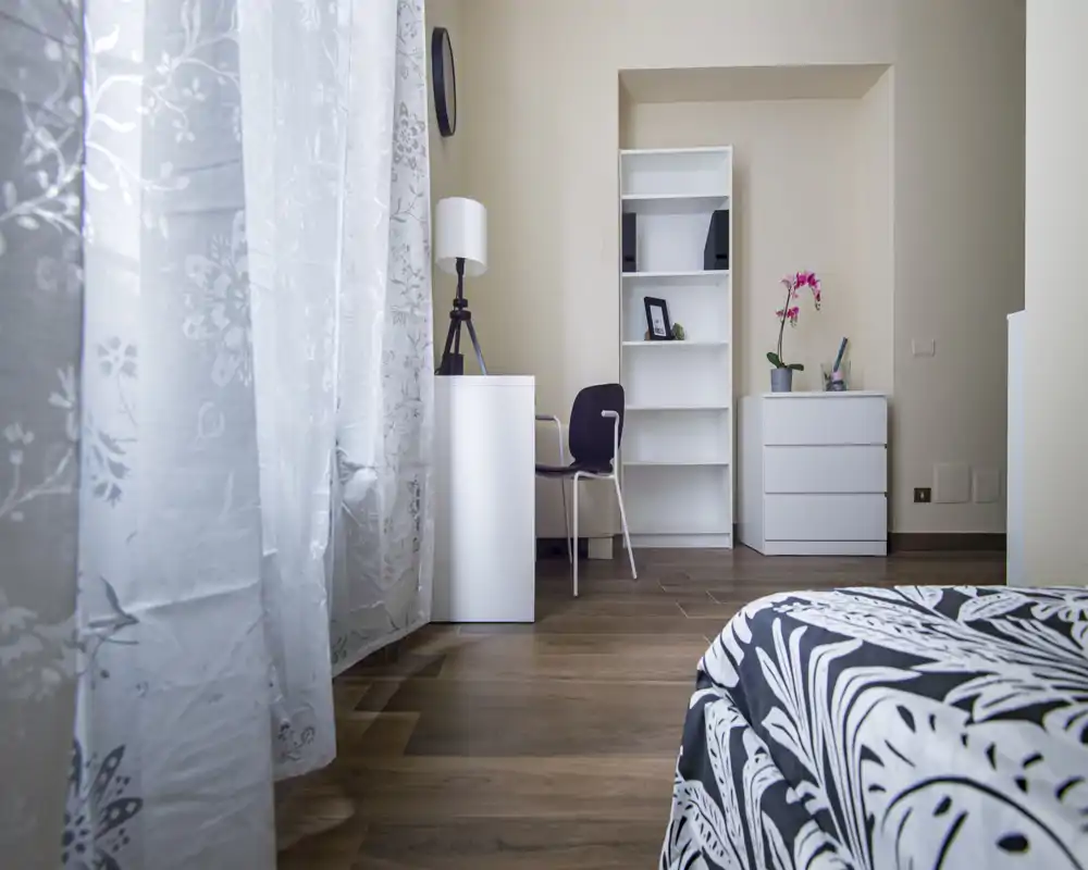 Angolo camera da letto moderno e luminoso con biancheria a fantasia, pavimento in legno e una piccola postazione con scaffale e cassetti — tende leggere lasciano entrare luce naturale.