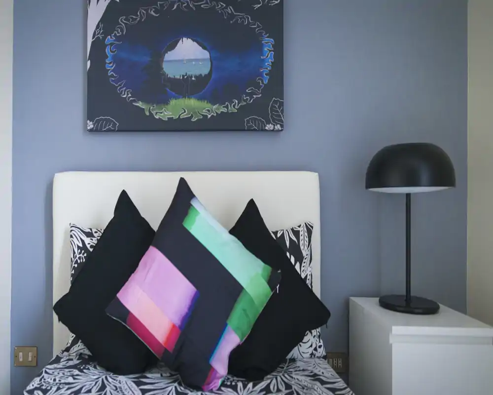 Angolo camera luminoso e curato con testata bianca, biancheria a fantasia e cuscini colorati, lampada da comodino moderna e quadro appeso.