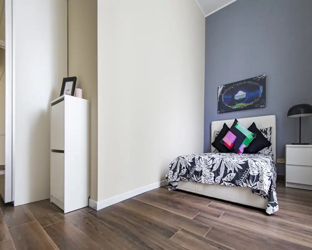 Camera da letto singola moderna e luminosa con copriletto a motivo, cuscini decorativi e lampada da comodino. Pavimento in legno e pareti neutre per un look pulito e contemporaneo.