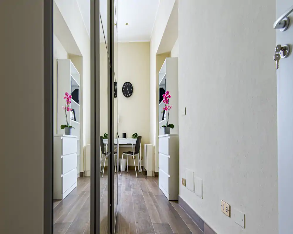 Corridoio luminoso e stretto con armadi a specchio e un piccolo angolo lavoro in fondo; pavimenti in legno moderno e orchidee decorative che danno calore.