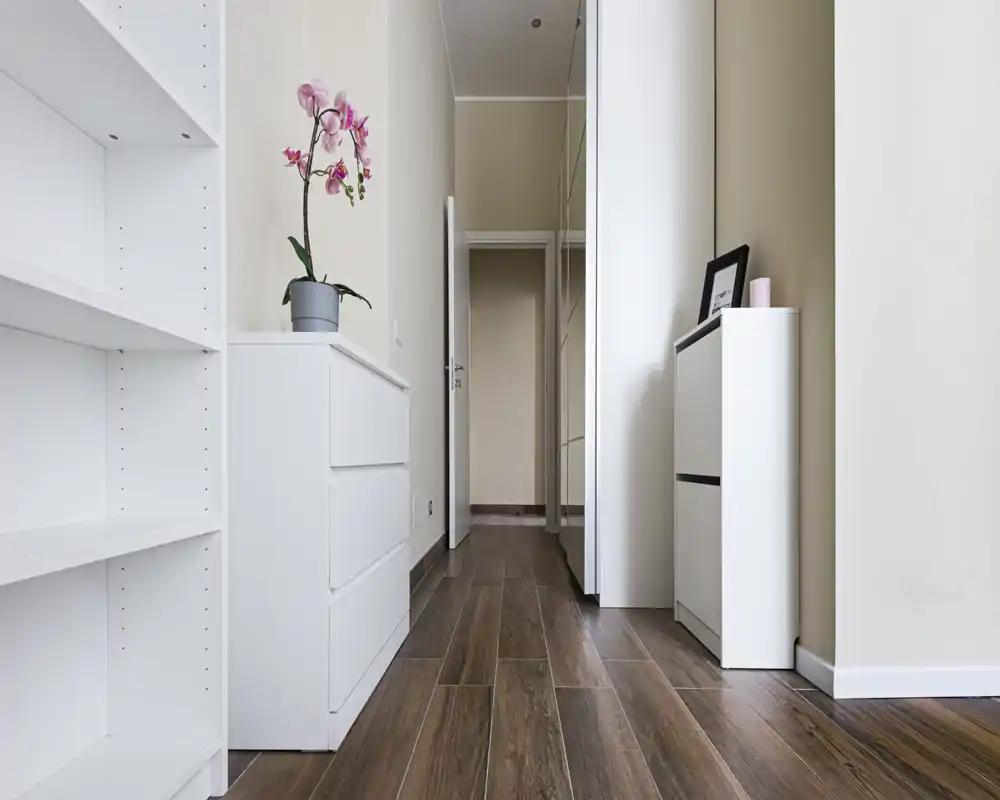 Ingresso/corridoio luminoso e minimale con pavimento effetto legno, mobili contenitore bianchi e un'orchidea decorativa — accogliente e ordinato.