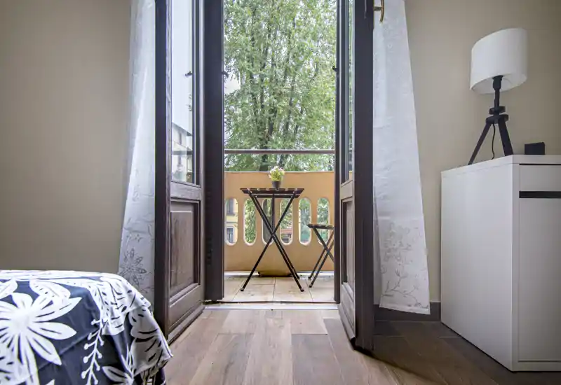 Camera da letto luminosa con accesso a un piccolo balcone e un tavolino bistro. Il letto ben rifatto, il comodino e la lampada creano un ambiente accogliente e ben illuminato, perfetto per gli annunci.