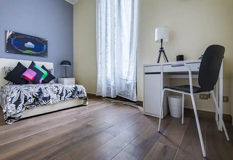 Camera da letto luminosa e ordinata con letto decorato, cuscini colorati e una scrivania vicino a una grande finestra — ideale per riposare e lavorare con luce naturale.
