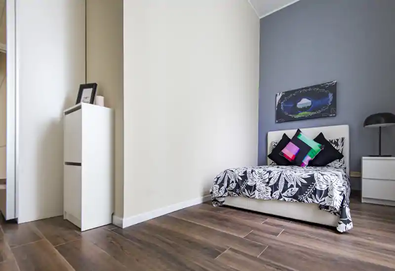 Camera da letto singola moderna e luminosa con copriletto a motivo, cuscini decorativi e lampada da comodino. Pavimento in legno e pareti neutre per un look pulito e contemporaneo.