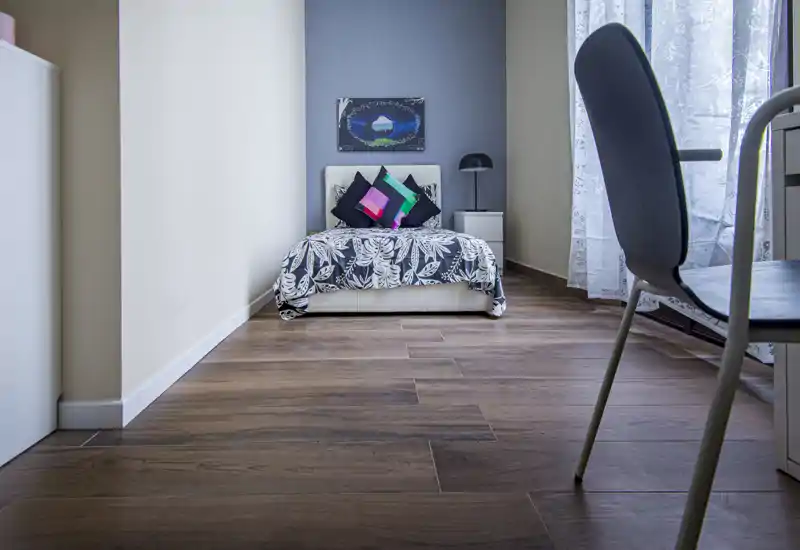 Camera da letto moderna e luminosa con un singolo letto ben rifatto, cuscini decorativi e una lampada da comodino accanto a una grande finestra che illumina la stanza.