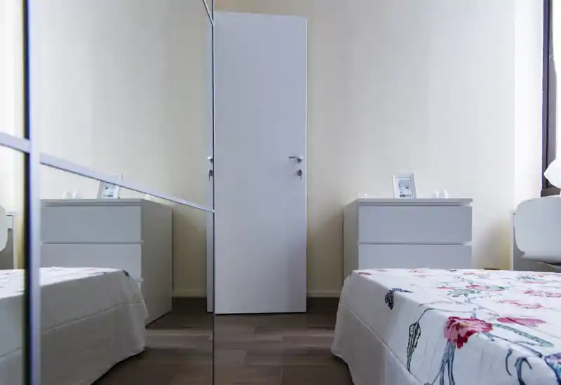 Camera da letto moderna e luminosa con copriletto floreale, comodini bianchi e armadio con specchio che amplia visivamente lo spazio.