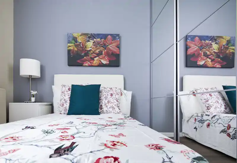 Camera da letto moderna e luminosa con copriletto decorato, cuscino accentato turchese, abat-jour e armadio con ante a specchio che crea profondità.