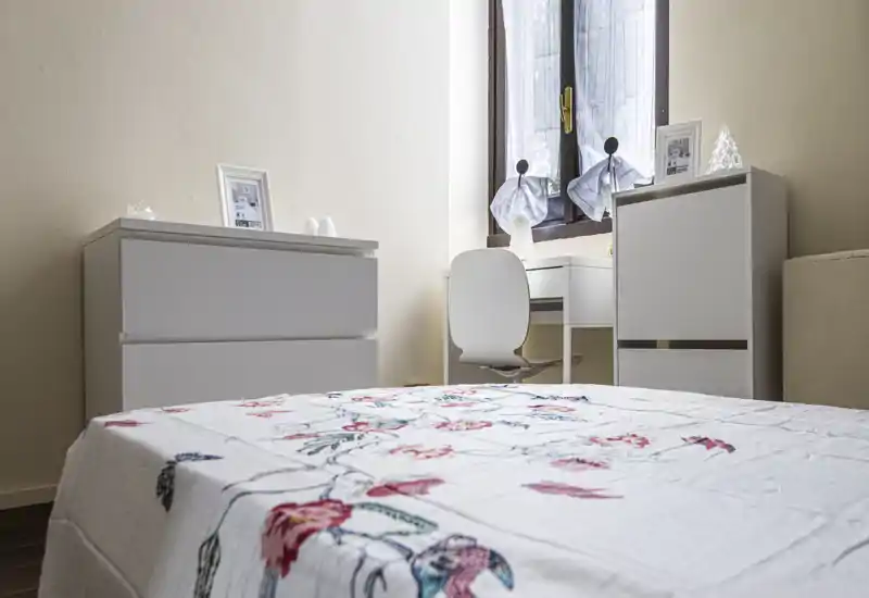 Camera da letto luminosa e ordinata con copriletto a fantasia in primo piano, due comò bianchi e una scrivania vicino alla finestra; toni neutri e luce naturale creano un'atmosfera rilassante.