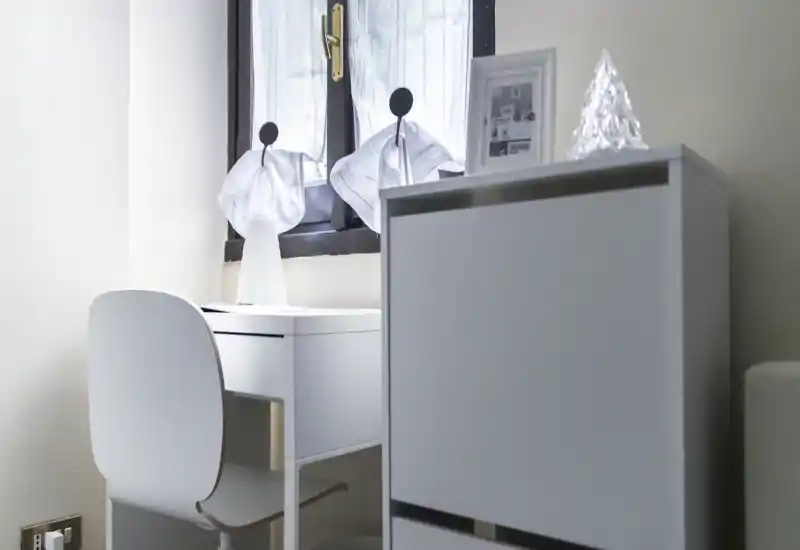 Minimalist bedroom corner with a small white desk and chair beside a window; bright, clean lines and subtle decorative items create a calm, functional atmosphere.