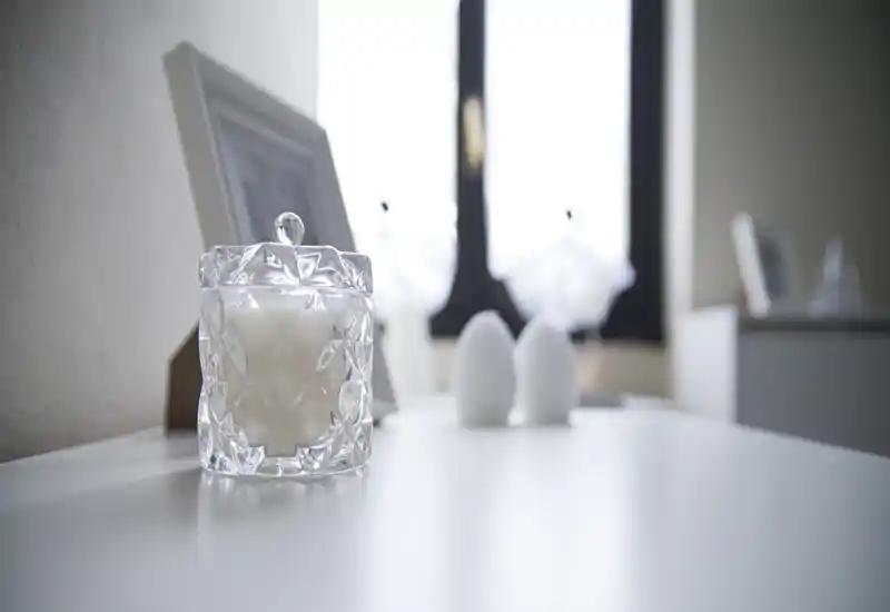 Close-up of decorative objects on a white surface: a faceted glass candle jar in the foreground with blurred home accessories and a window in the background. The image emphasizes styling detail rather than showing a full room.