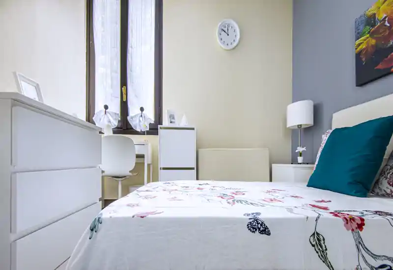 Camera da letto luminosa e moderna con copriletto floreale, cuscino accentato turchese e comodino ordinato con lampada; luce naturale da una finestra alta che crea un'atmosfera fresca.