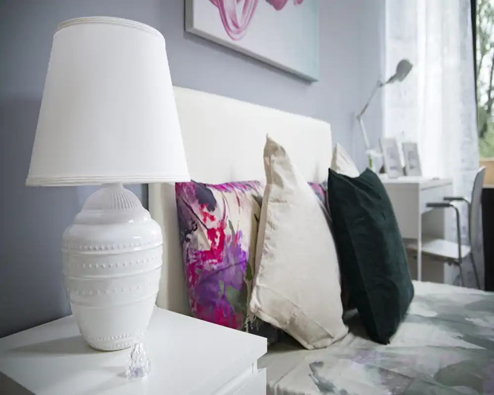 Bright, styled bedroom close-up with decorative pillows, a white headboard and bedside lamp — ideal for listing photos highlighting comfort and decor.