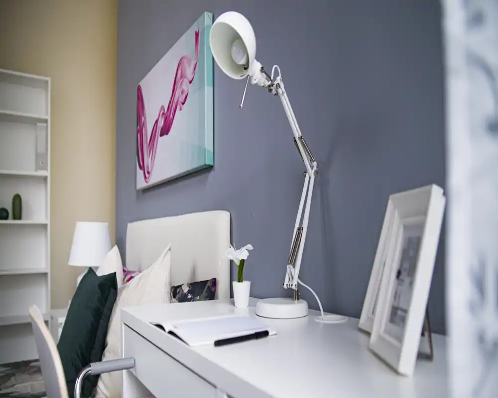 Contemporary bedroom corner with a white desk and articulated lamp beside a padded headboard, decorative pillows and wall art; bright, styled scene ideal for property listings.