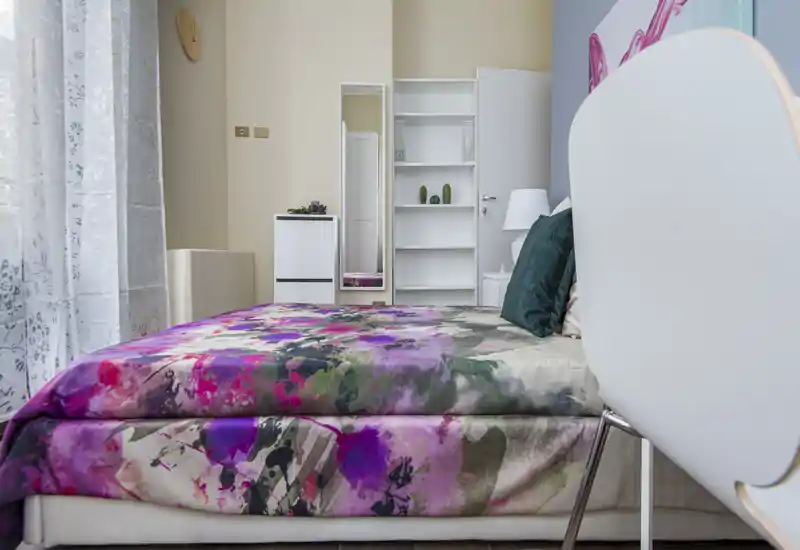 Bright, modern bedroom with a colorful floral duvet, visible mattress and pillows, bedside lamp and shelving — well-lit by a large window.
