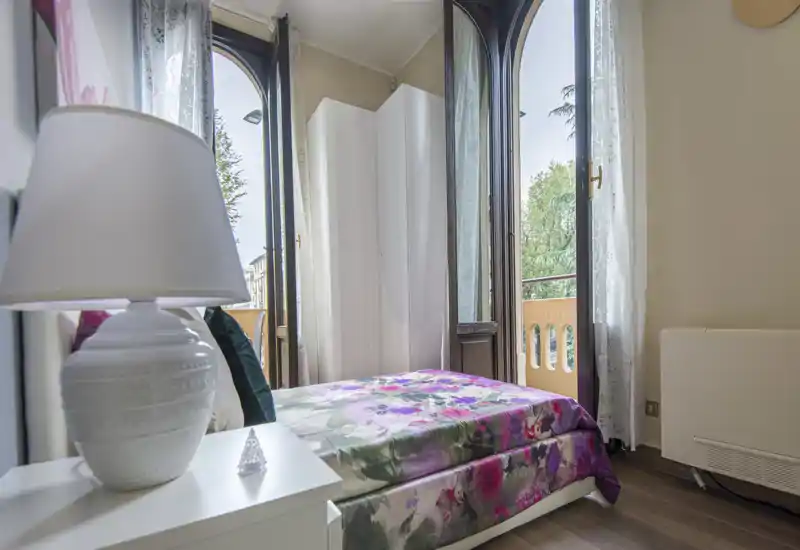 Sunny, cozy bedroom with a floral bedspread, bedside lamp and tall arched windows opening to a small balcony — bright and welcoming space ideal for a rental listing.