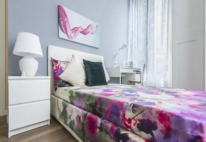 Stylish single bedroom with colorful floral bedding, decorative pillows and a bright work desk by a sheer-curtained window — ideal for a cozy, modern rental.