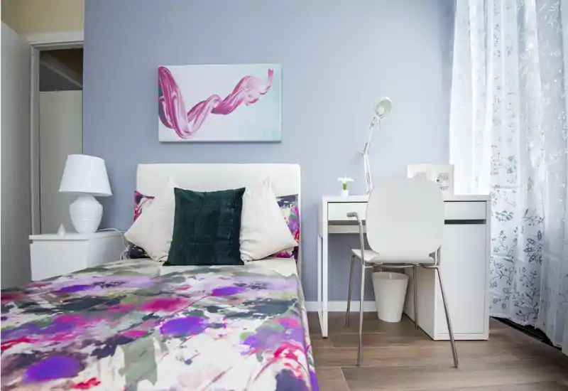 Stylish bedroom with a floral duvet, plush pillows and a compact white desk by the window. Bright natural light and coordinated decor create a fresh, welcoming atmosphere ideal for short-term rentals.