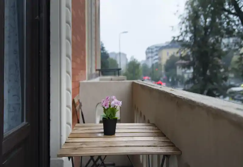 Cozy small balcony with a wooden bistro table and a potted pink flower, overlooking a leafy urban street — ideal for morning coffee.