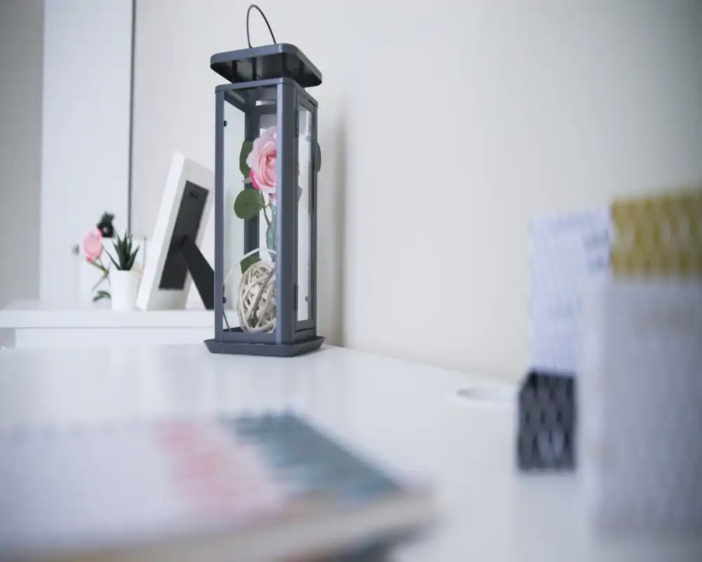 Angolo soggiorno luminoso e minimale evidenziato da elementi decorativi: una lanterna con rosa artificiale, piccole piante e cornici su una superficie bianca ordinata, perfetto per un ambiente moderno e tranquillo.
