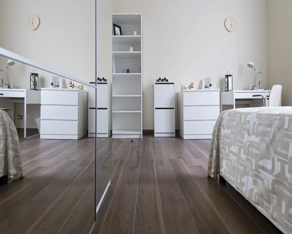 Camera da letto luminosa e moderna con mobili bianchi minimal, lampade decorative sui comodini e un letto confortevole in primo piano. Lo spazio appare arioso grazie alla luce naturale e al pavimento in legno caldo.