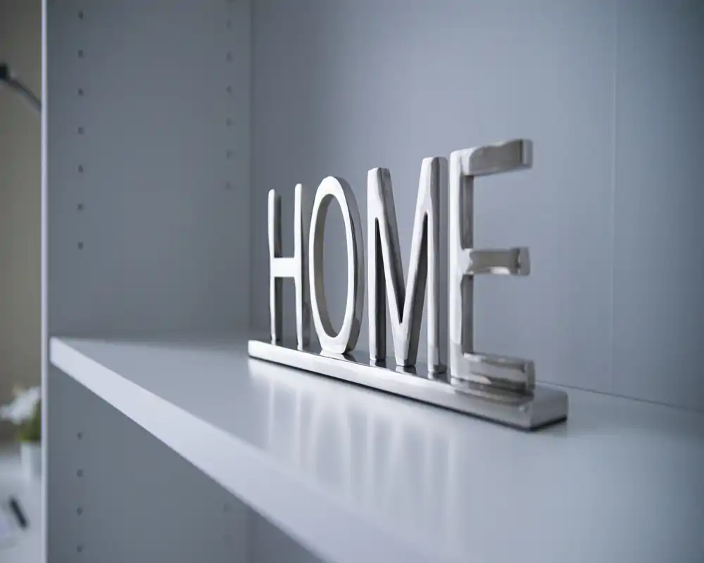 Primer plano de un adorno metálico con la palabra "HOME" sobre una estantería blanca; escasos elementos contextuales y fondo neutro.