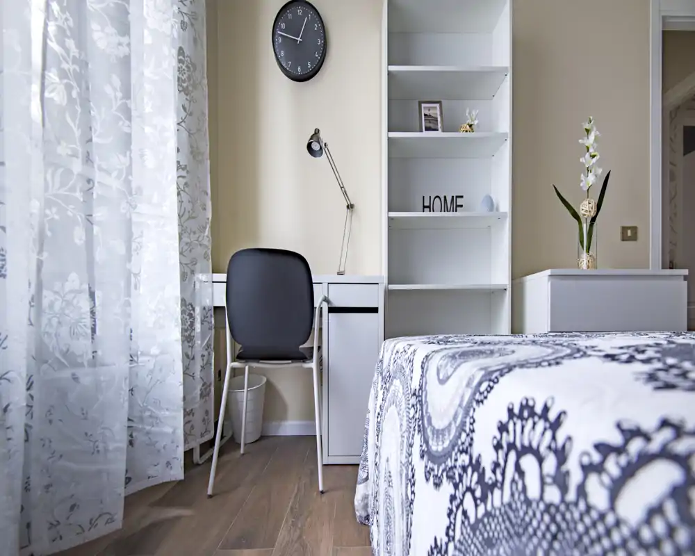 Dormitorio luminoso y bien decorado con colcha estampada, escritorio y estanterías; la luz natural a través de la cortina ligera genera un ambiente acogedor.
