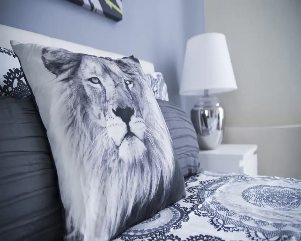Detalle de dormitorio con un cojín decorativo con estampado de león sobre una cama bien hecha, ropa de cama estampada y una lámpara de noche que crean un ambiente moderno y tranquilo.