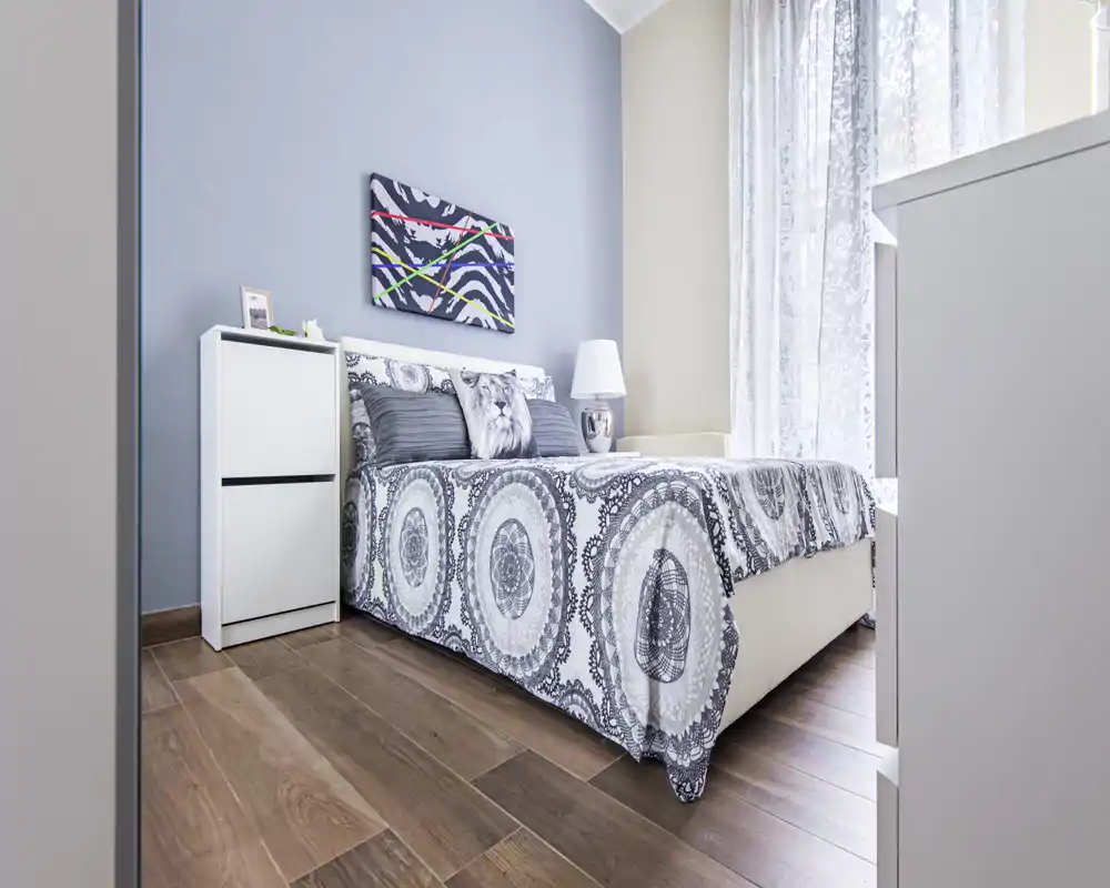 Dormitorio luminoso y bien presentado con edredón estampado, cojines decorativos y luz natural suave desde la ventana — ideal para fotos de alquiler.
