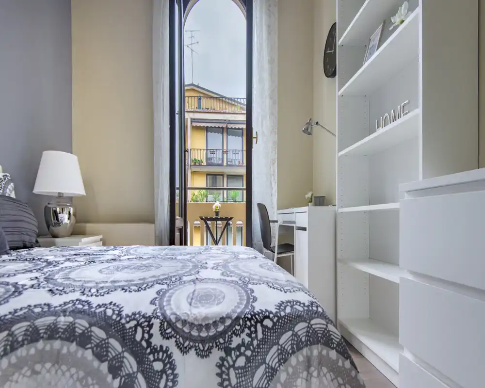 Dormitorio luminoso y bien decorado con ropa de cama estampada, lámpara de noche y escritorio junto a una gran puerta balcón arqueada que se abre al exterior.