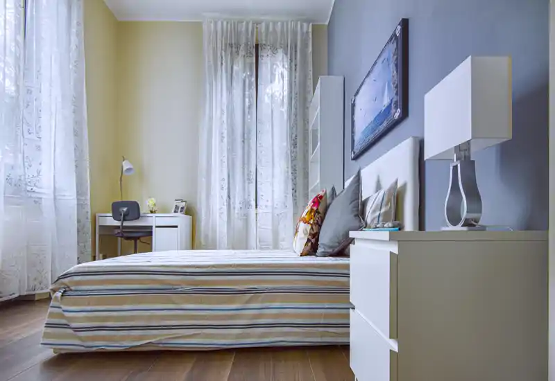 Camera da letto luminosa e curata, con letto basso, cuscini decorativi e tende leggere che diffondono luce naturale.