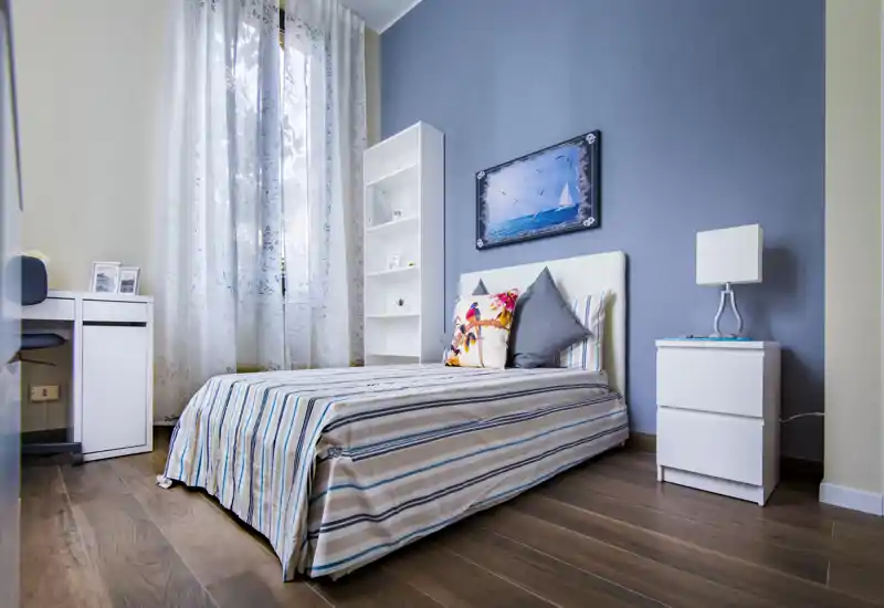 Camera da letto moderna e luminosa con letto a righe, cuscini decorativi e comodino — ben arredata e illuminata da una finestra alta.