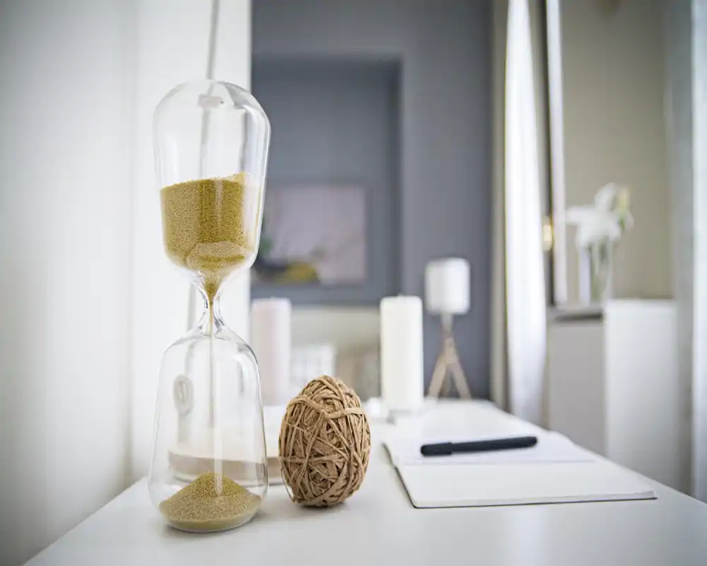 Primer plano de una superficie del área de estar con objetos decorativos: un reloj de arena, una bola tejida y un bloc de notas, que sugieren una mesa consola acogedora en un salón con luz suave.