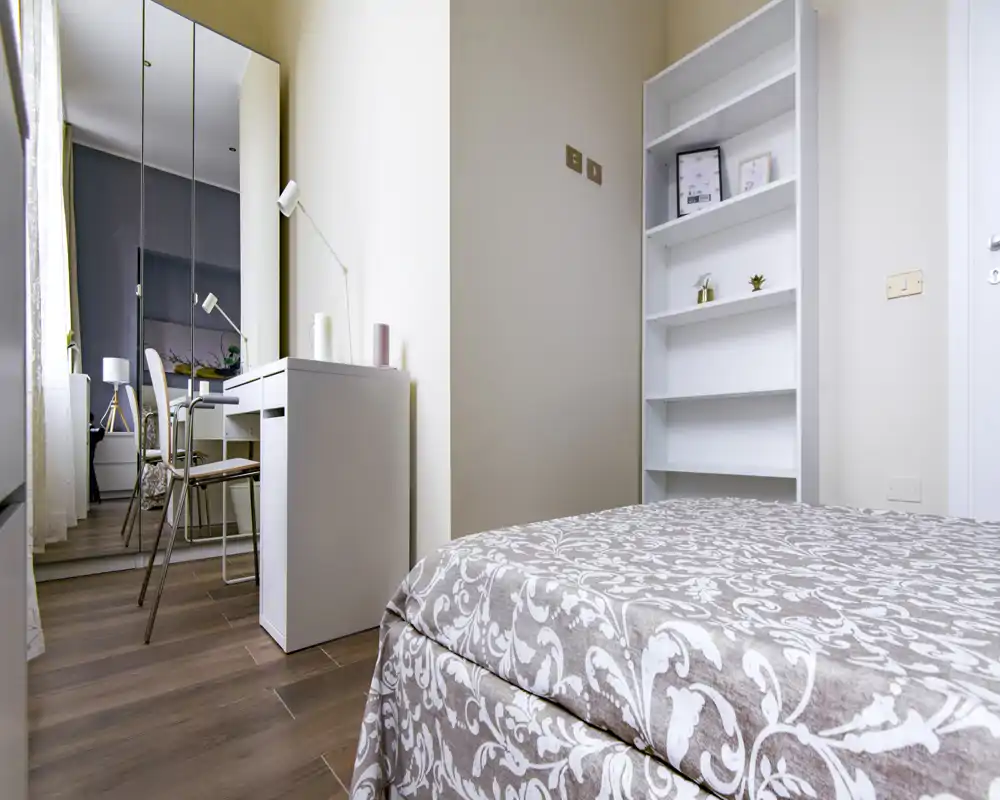 Rincón de dormitorio luminoso y ordenado con colcha estampada, estantería blanca y un pequeño escritorio con espejo que refleja más detalles de la habitación.