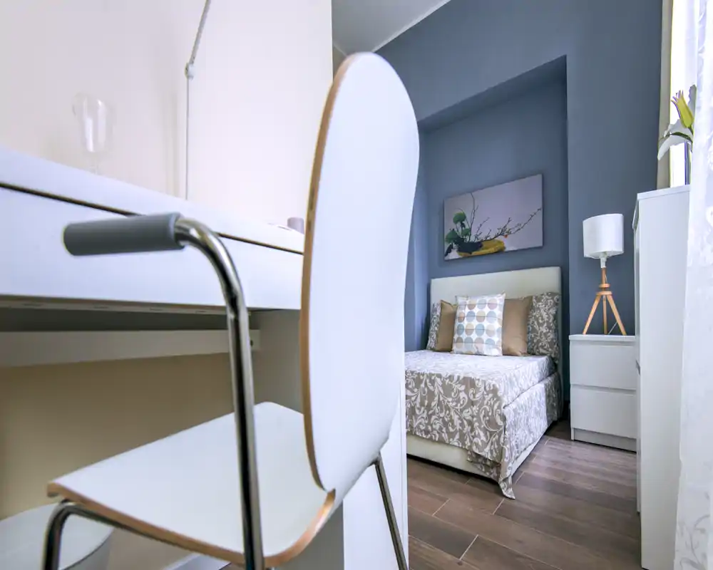 Dormitorio individual luminoso y bien decorado con colcha estampada, cojines decorativos y lámpara de mesita; en primer plano se ve un pequeño espacio de trabajo con silla.