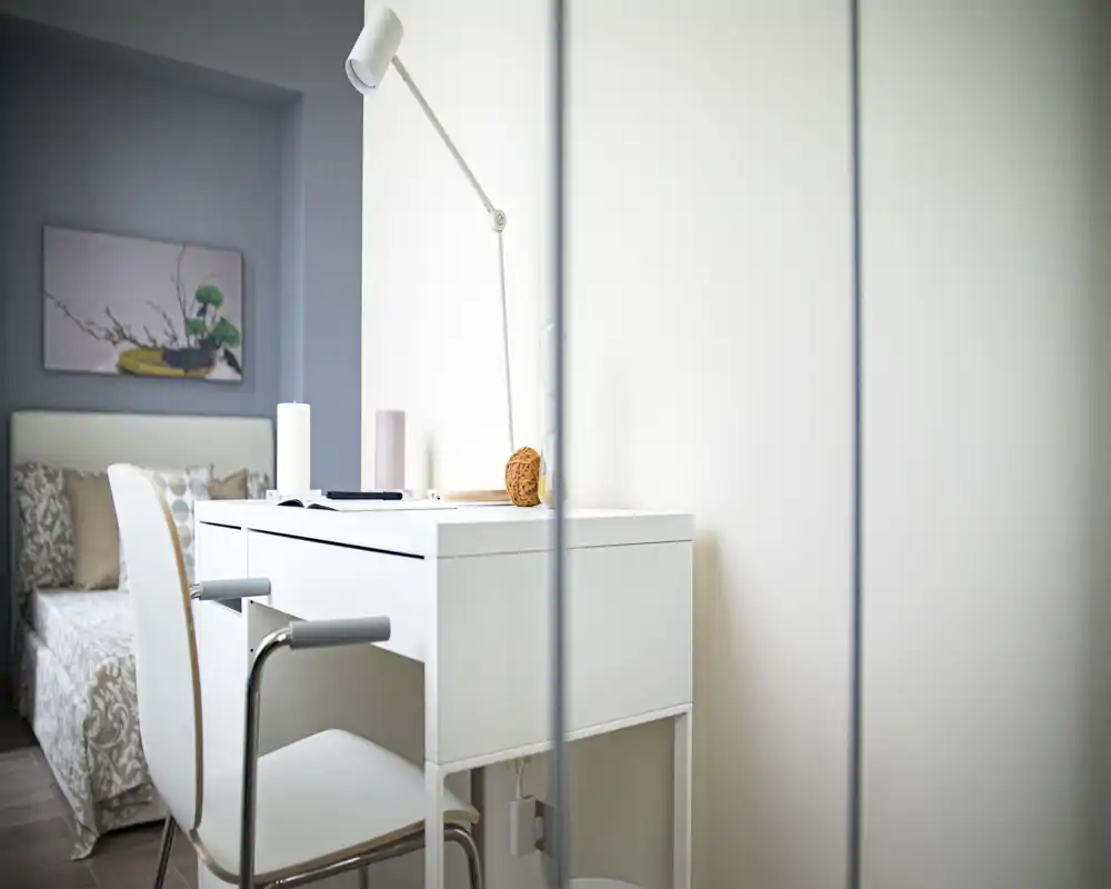 Rincón de dormitorio luminoso y minimalista con escritorio blanco y silla en primer plano y cama parcialmente visible con cojines al fondo. Estilo limpio y moderno con textiles suaves y lámpara de trabajo, perfecto para estudios o habitaciones de invitados.