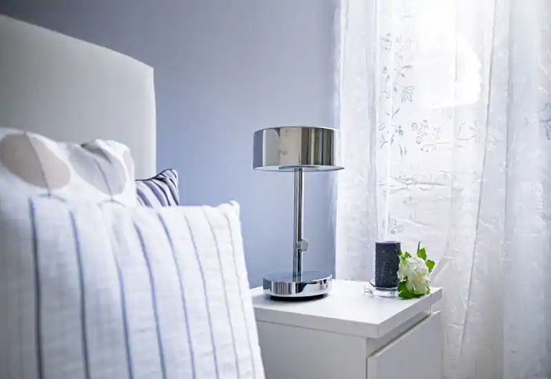 Angolo camera luminoso e sereno con cuscini a righe, una moderna lampada da comodino cromata e tende leggere floreali che diffondono luce naturale.