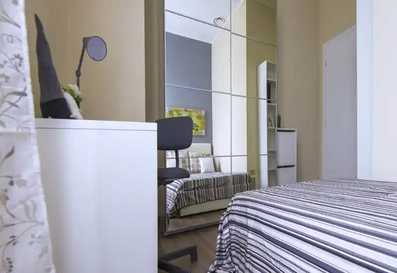 Bright, modern bedroom with a striped bedspread, mirrored wardrobe creating depth, and a small desk and chair—well-lit and tastefully furnished for short stays.