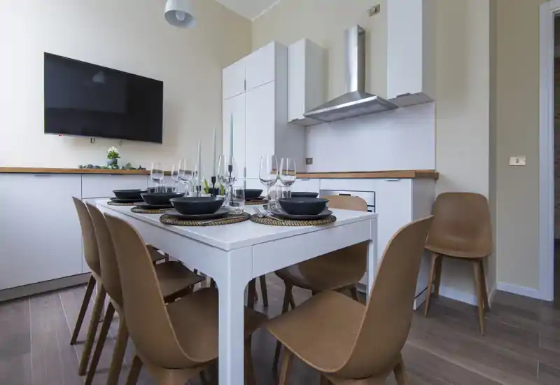 Modern dining area integrated with a compact kitchen: a white table set for six with elegant place settings, warm brown chairs and a minimalist kitchen backdrop — ideal for apartment listings.