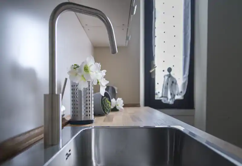 Bright modern kitchen sink with a stainless-steel tap and decorative flowers on a warm wooden countertop, lit by natural window light.