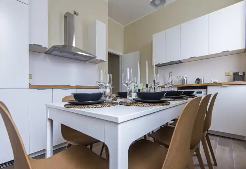 Stylish dining area integrated with a modern kitchen. A white table is set for several guests with elegant glassware and place settings, paired with warm-toned chairs and minimal cabinetry.