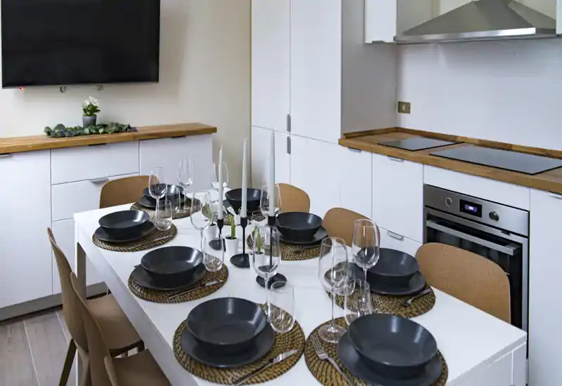 A modern, well-staged dining area integrated with the kitchen, featuring a white table set for six with woven placemats, dark bowls and glassware — ideal for showcasing apartment dining space.