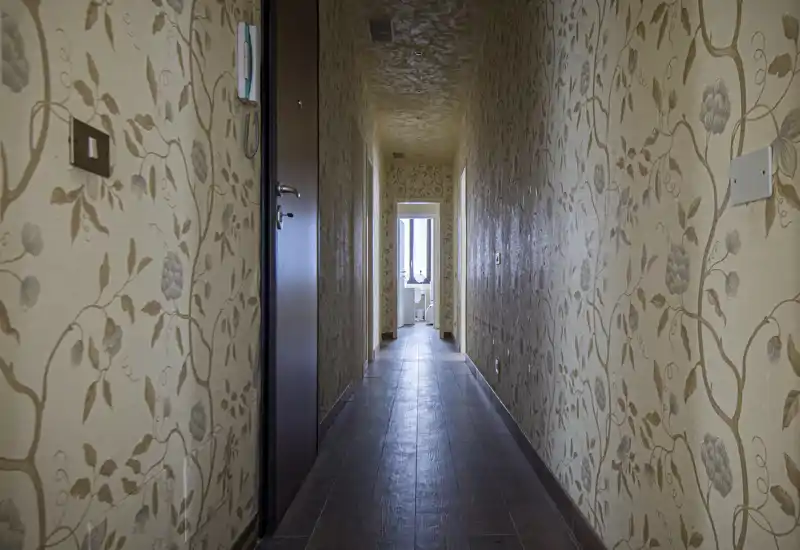 Long interior hallway with patterned wallpaper, wooden-look floor and door on the left; natural light visible from a room at the far end.