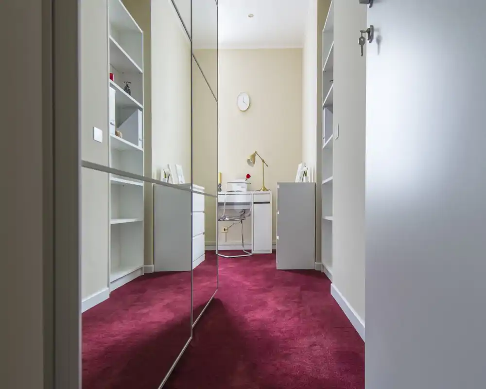 Small dressing area or corridor with mirrored wardrobe doors, built-in shelving and a compact desk at the far end; plush red carpet and soft neutral walls create a warm contrast.